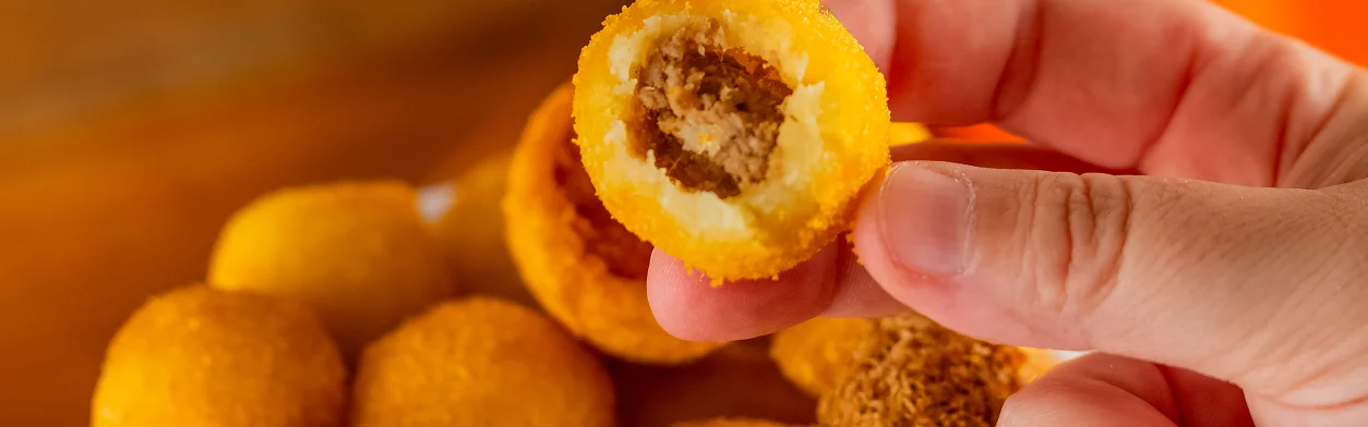 Mão segurando uma coxinha de carne mordida. No fundo uma tigela de salgados - Vó Linda Casa De Salgados
