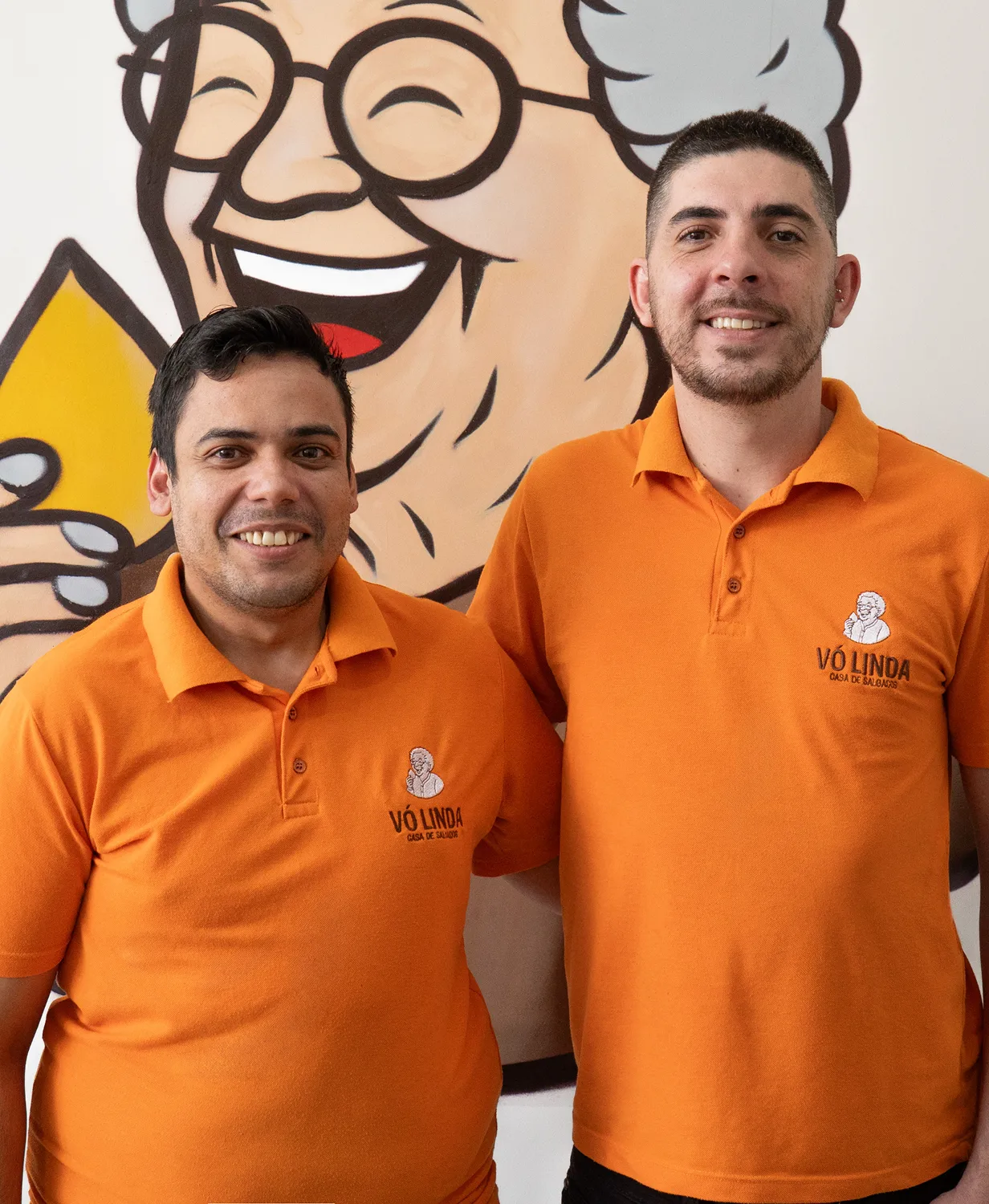 Fundadores da Vó Linda Casa de Salgados posando com uniforme laranja em frente ao mural da marca